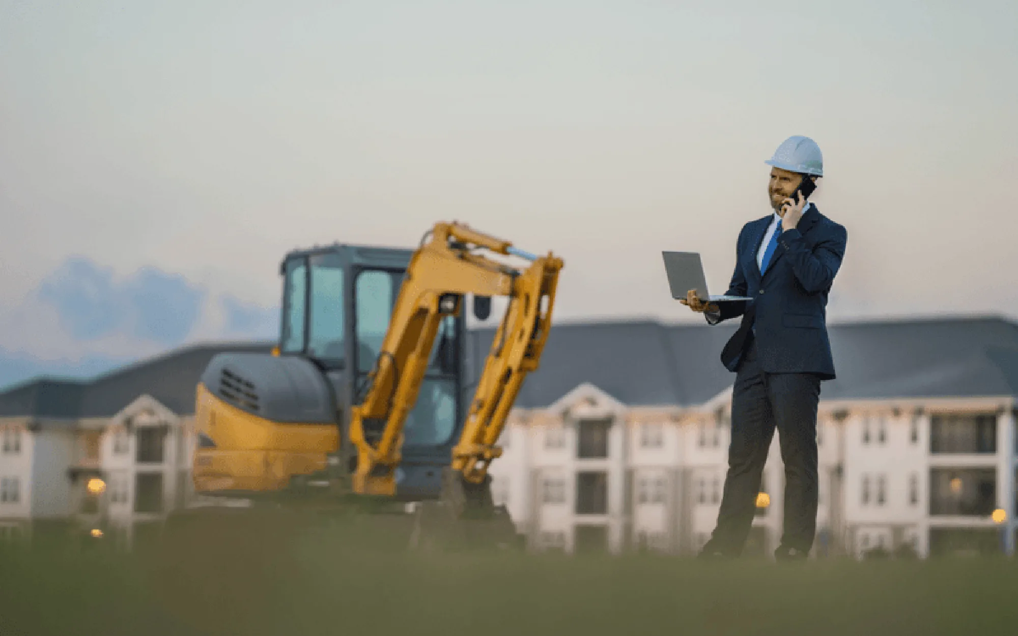 Ein Bauleiter steht auf einer Baustelle neben einem Bagger, telefoniert und arbeitet gleichzeitig mit einem Laptop.