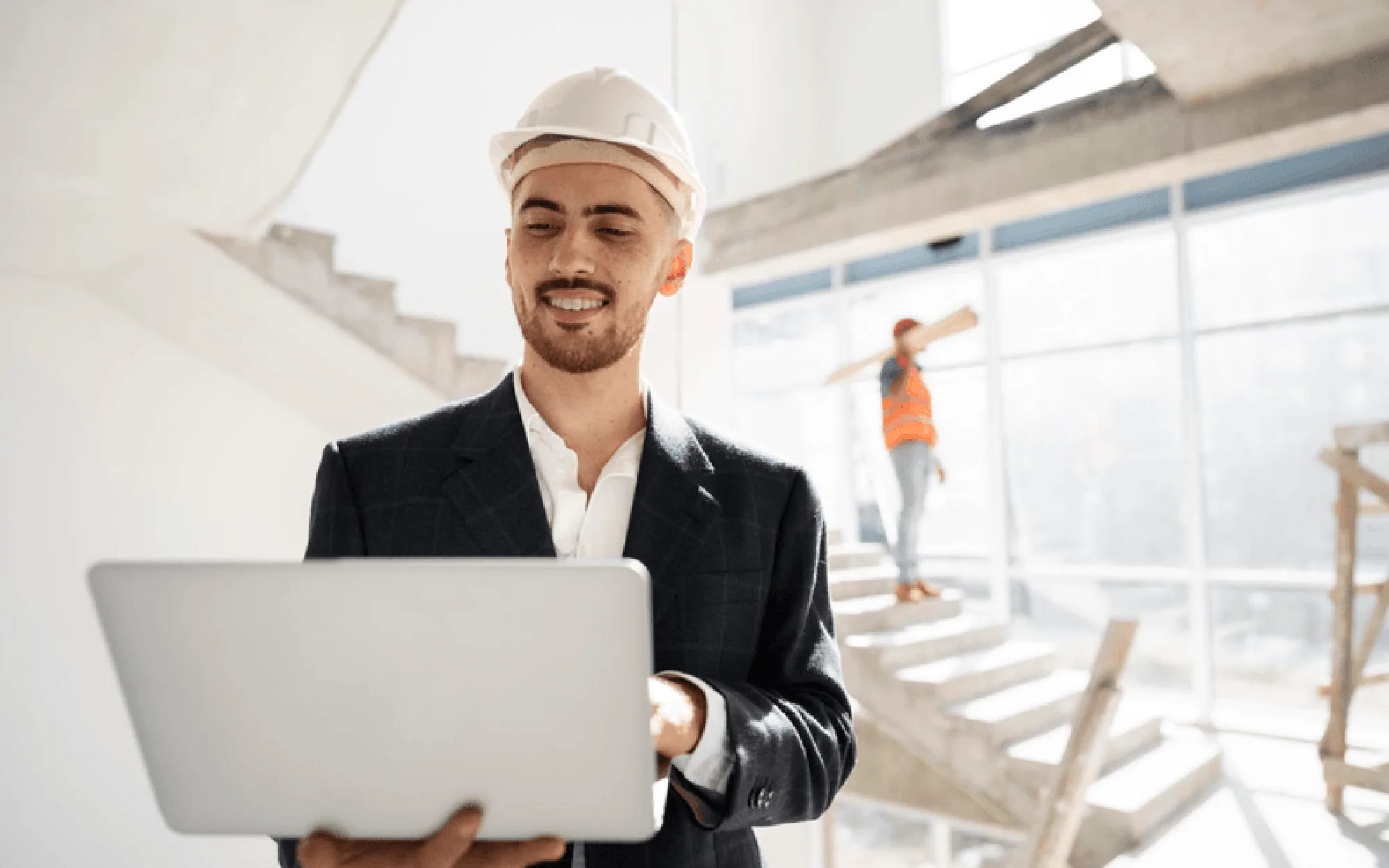 Ein Architekt trägt einen Schutzhelm und arbeitet auf einer Baustelle mit einem Laptop, während im Hintergrund ein Bauarbeiter zu sehen ist.